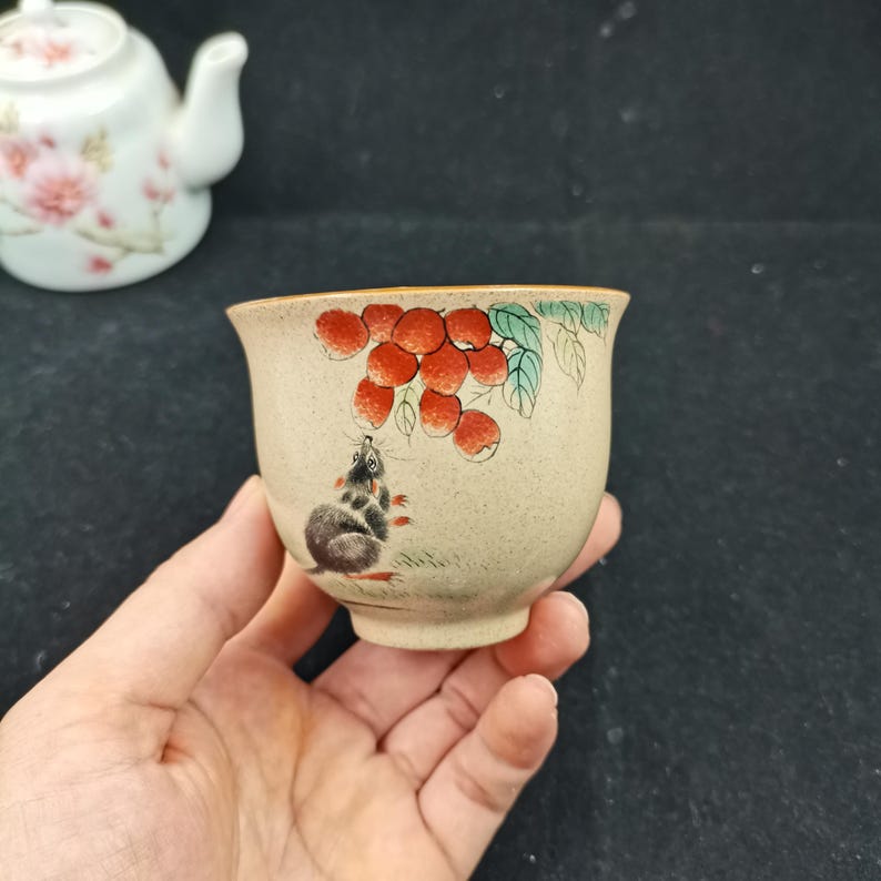 May include: A small, speckled beige ceramic tea cup featuring a hand-painted illustration of a grey mouse looking up at a cluster of red lychee fruits with green leaves. The cup has a flared rim and a smooth, rounded base, showcasing detailed artwork.