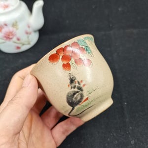 May include: A beige ceramic tea cup featuring a hand-painted illustration of a small gray mouse reaching toward a cluster of red lychee fruits with green leaves. The cup has a speckled texture and a smooth, rounded shape with a flared rim.