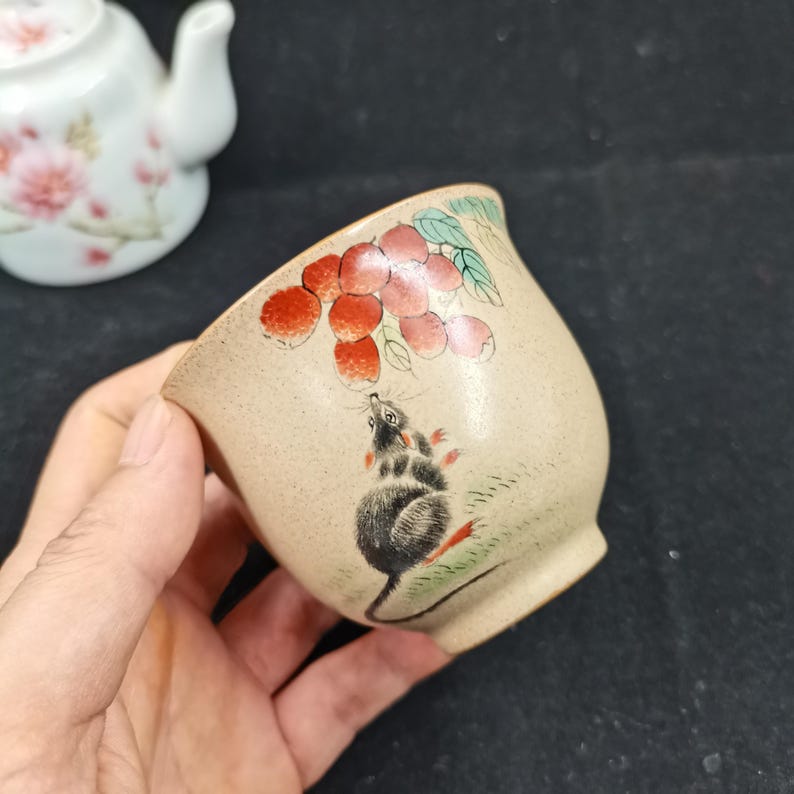 May include: A beige ceramic tea cup featuring a hand-painted illustration of a small gray mouse reaching toward a cluster of red lychee fruits with green leaves. The cup has a speckled texture and a smooth, rounded shape with a flared rim.
