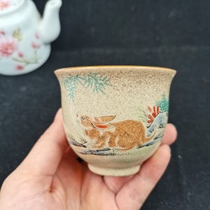 May include: A ceramic tea cup with a speckled beige finish featuring a hand-painted brown rabbit, green pine needles, and red floral accents. The cup has a flared rim and a rounded body, with a white teapot visible in the blurred background.
