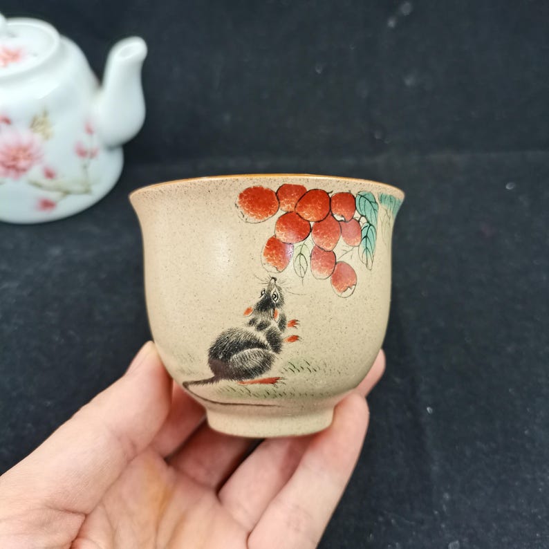 May include: A small, speckled beige ceramic tea cup featuring a hand-painted illustration of a dark gray mouse looking up at a cluster of red lychee fruits with green leaves. The cup has a smooth, rounded rim and a tapered base design.
