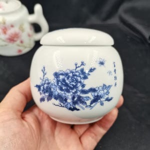 Puede incluir: Un tarro de cerámica blanca con tapa, que presenta un patrón floral azul de peonías y pequeñas mariposas. El diseño incluye un delicado trabajo de pincel en tinta azul y caracteres chinos en el lateral del recipiente.