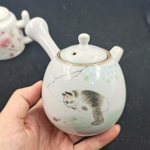 Op de afbeelding: Een witte keramische theepot met een zijhandvat en een deksel, voorzien van een geschilderde illustratie van een pluizige kat en een kleine slak. De theepot heeft een craquelé glazuurafwerking en delicate bloemaccenten in pasteltinten.