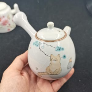 Puede incluir: Una tetera de cerámica blanca con asa lateral y tapa, que presenta una ilustración pintada de un gato naranja claro sentado bajo una rama de pino. La tetera tiene un acabado de esmalte craquelado sutil y un pequeño pico vertedor.