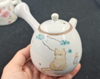 Pode incluir: Uma chaleira de cerâmica branca com pega lateral e tampa, apresentando uma ilustração pintada de um gato laranja claro sentado sob um ramo de pinheiro. A chaleira tem um acabamento de esmalte craquelado subtil e um pequeno bico.