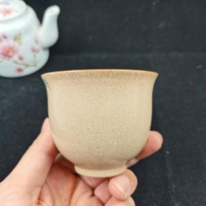 May include: A small, speckled beige ceramic tea cup with a smooth, rounded rim and a tapered base. The cup features a subtle, light brown speckled texture throughout its exterior surface, providing a neutral and earthy appearance for tea service.