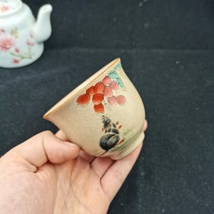 May include: A small, speckled beige ceramic tea cup featuring a hand-painted illustration of a grey mouse reaching toward a cluster of red berries with green leaves. The cup has a flared rim and a smooth, rounded body, showcasing detailed brushwork.
