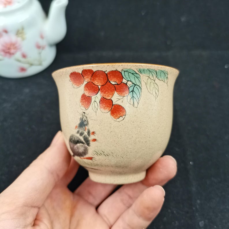 May include: A beige ceramic tea cup with a speckled finish featuring a hand-painted illustration of a small grey mouse looking up at a cluster of red berries with green leaves. The cup has a flared rim and a small base, with a matching teapot visible.