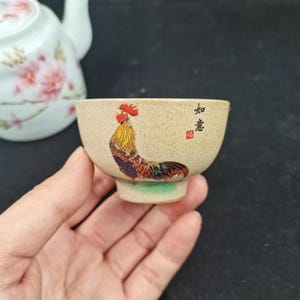 Könnte beinhalten: Eine kleine, gesprenkelte beige Keramik-Teetasse mit einer farbenfrohen Hahn-Illustration auf der Vorderseite. Das Design enthält schwarze chinesische Kalligrafiezeichen und einen kleinen roten quadratischen Stempel. Eine passende weiße Teekanne ist sichtbar.