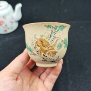 Può includere: Una tazza da tè in ceramica beige con una consistenza maculata, caratterizzata da un'illustrazione dipinta a mano di una scimmia marrone che si arrampica su un pino verde. La tazza ha un bordo svasato e un corpo arrotondato con dettagli marroni e verdi.