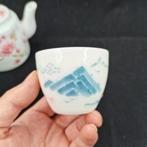 May include: A small white ceramic tea cup featuring a hand-painted blue mountain landscape design. The cup has a smooth, rounded shape and includes a small red stamp mark on the side. A matching white teapot with floral patterns is visible in the background.