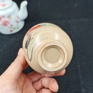 May include: A beige ceramic cup with a speckled texture and a hand-painted floral design featuring red and green accents. The cup has a circular base and is positioned near a white teapot decorated with pink flowers and green leaves.