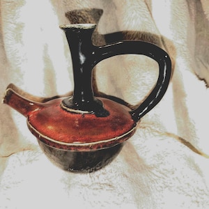 May include: A ceramic vessel featuring a rounded base and a wide, flat shoulder finished in a glossy reddish-brown glaze. It has a tall, slender black neck, a curved black handle, and a short, angled spout extending from the side of the body.