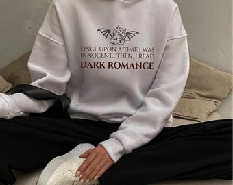 May include: A white hooded sweatshirt featuring a black graphic of a winged cherub above the text "ONCE UPON A TIME I WAS INNOCENT... THEN, I READ DARK ROMANCE" with the words "DARK ROMANCE" printed in a dark red color on the chest.
