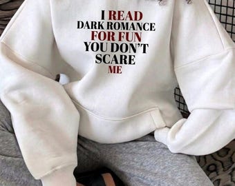 May include: A white hooded sweatshirt featuring the text I READ DARK ROMANCE FOR FUN YOU DON'T SCARE ME printed in black and dark red lettering. The garment has long sleeves with ribbed cuffs, a hood, and a front kangaroo pocket.