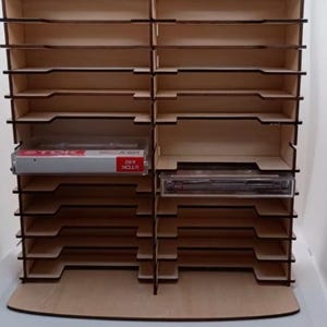 May include: A light-colored wooden storage rack with two vertical columns of horizontal shelves designed for cassette tapes. One TDK A60 cassette case is visible on the left side. The unit features a flat base and a separate detached wooden piece.