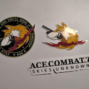 May include: Three vinyl stickers featuring Ace Combat 7 imagery. One circular black sticker displays a yellow wolf emblem with the text TRIGGER AND YOU'LL MAKE IT THROUGH, STUCK WITH TRIGGER, and 444. A second wolf sticker and a white ACE COMBAT 7 SKIES UNKNOWN logo sticker.