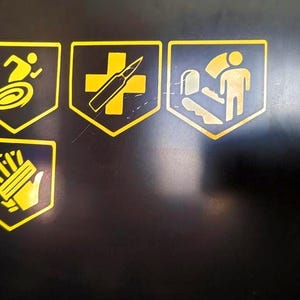 May include: A black surface featuring four yellow shield-shaped decals. The top row displays a running figure over a spiral, a bullet over a cross, and a figure standing by a staircase. The bottom row shows a hand with multiple small rectangular shapes.