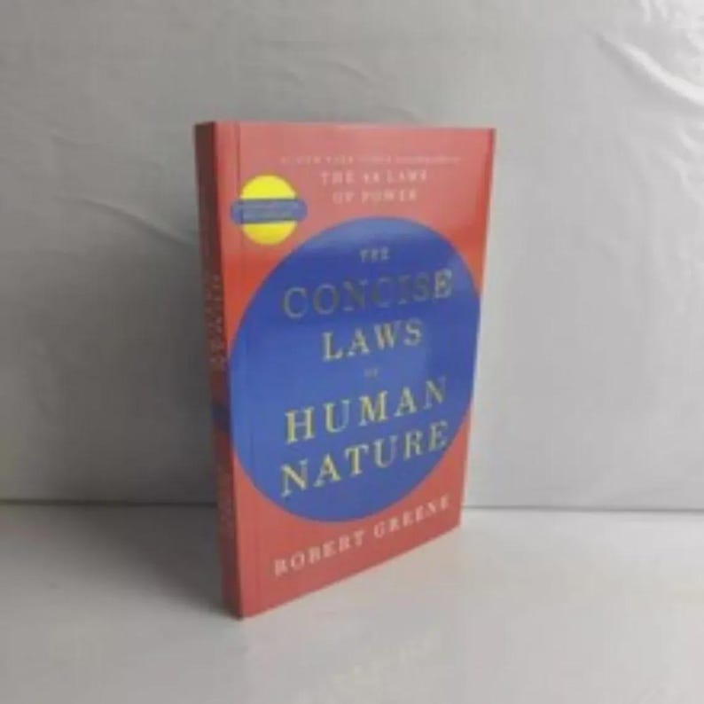Pu&ograve; includere: Un libro con copertina rigida intitolato The Concise Laws of Human Nature di Robert Greene. La copertina presenta un grande cerchio blu su sfondo rosso con testo dorato. Un adesivo circolare giallo &egrave; visibile sul lato superiore sinistro della copertina.