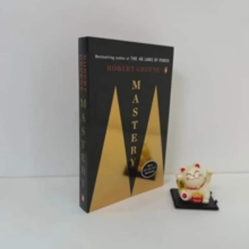 Pu&ograve; includere: Un libro nero e oro intitolato Mastery di Robert Greene, caratterizzato da un design geometrico triangolare sulla copertina, posizionato accanto a una piccola statuetta di gatto portafortuna bianco con dettagli rossi su base nera.