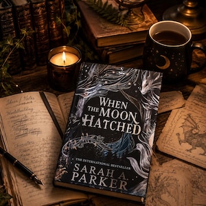 Pode incluir: Um livro de capa dura intitulado When the Moon Hatched de Sarah A. Parker com uma capa escura ilustrada apresentando um dragão, sobre uma superfície de madeira ao lado de um caderno aberto com notas manuscritas e uma caneta-tinteiro.