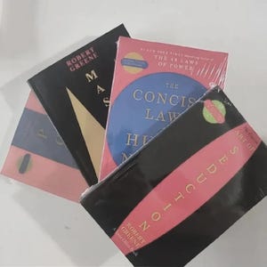 Pu&ograve; includere: Una collezione di tre libri di Robert Greene, tra cui The Concise Laws of Human Nature con un cerchio blu su copertina rosa, Mastery con copertina nera e triangolo dorato, e The Art of Seduction con un design nei colori nero e rosa.