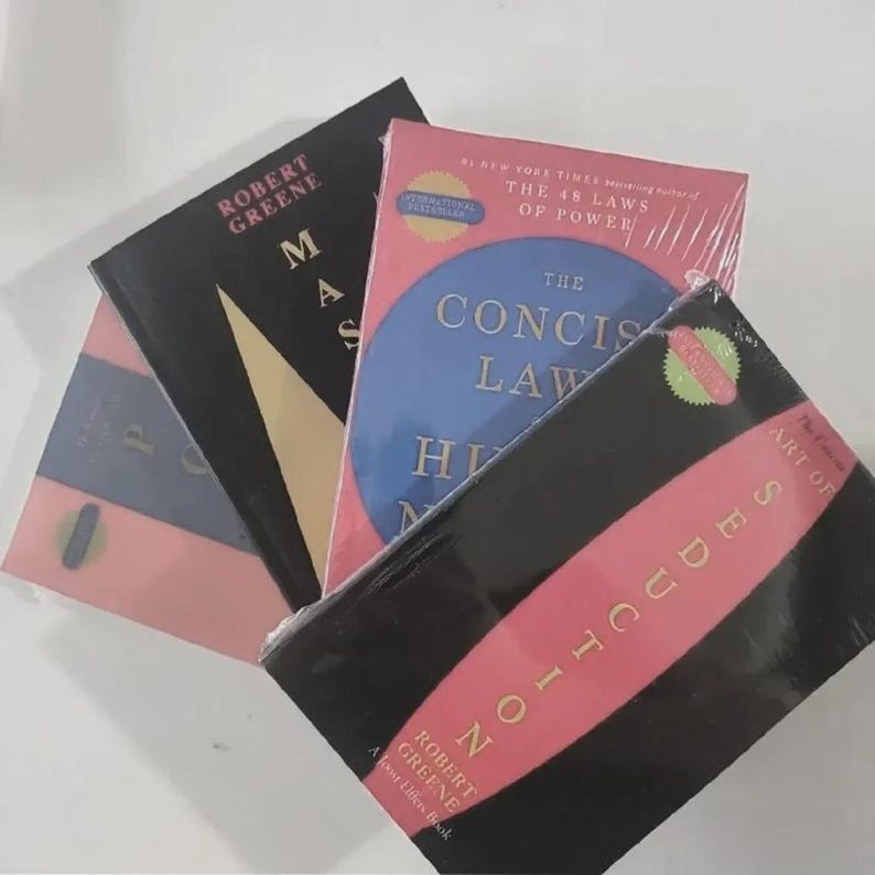 Pu&ograve; includere: Una collezione di tre libri di Robert Greene, tra cui The Concise Laws of Human Nature con un cerchio blu su copertina rosa, Mastery con copertina nera e triangolo dorato, e The Art of Seduction con un design nei colori nero e rosa.