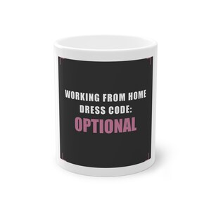 May include: A white ceramic mug featuring a black rectangular graphic on the front. The text reads "WORKING FROM HOME DRESS CODE: OPTIONAL" in white and pink capital letters, framed by thin pink corner accents on the black background.