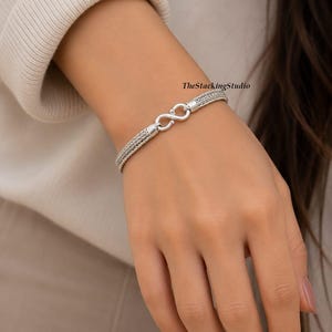 Può includere: Un bracciale color argento caratterizzato da un design a catena intrecciata a doppio filo collegato da una chiusura a forma di infinito. Il testo TheStackingStudio è visibile vicino alla chiusura. Il bracciale ha una finitura metallica lucida.
