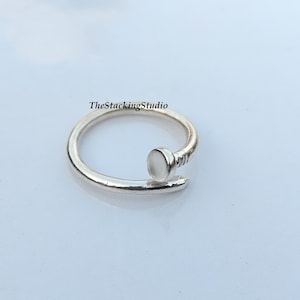 Könnte beinhalten: Ein silberfarbener Ring in Form eines gebogenen Nagels mit flachem, rundem Kopf und spitz zulaufendem Ende. Das Band hat eine glatte, polierte Oberfläche und einen kleinen texturierten Bereich nahe dem Kopf. Der Text TheStackingStudio ist zu sehen.