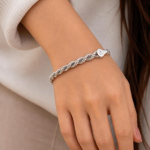 Può includere: Un bracciale a catena a corda color argento con un design in metallo intrecciato. Il bracciale include un piccolo componente di chiusura piatto a forma di goccia, chiaramente stampigliato con il numero 925, che indica la composizione in argento.