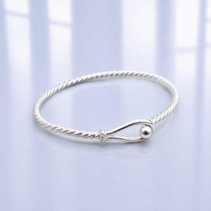 Puede incluir: Un brazalete de color plateado con una textura trenzada similar a una cuerda a lo largo de toda la banda. El brazalete termina en un mecanismo de cierre tipo gancho, que incluye una pequeña cuenta esférica y lisa en el extremo.