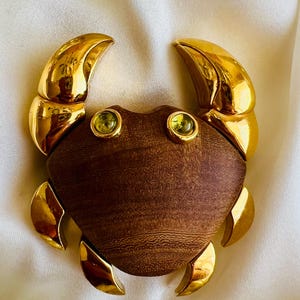 May include: A crab-shaped brooch featuring a polished gold-tone metal body with two large upper claws and four smaller lower legs. The central shell is crafted from dark brown wood, accented by two small, round, light green faceted gemstone eyes.
