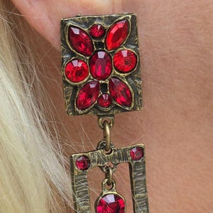 May include: A drop earring featuring an antique brass-toned metal frame with a textured finish. The top square section is adorned with faceted red crystals in a floral pattern, followed by a rectangular frame and a dangling teardrop-shaped red crystal.