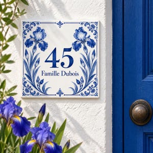 Könnte beinhalten: Ein quadratisches, weißes Keramik-Hausnummernschild mit einem blauen Blumenmuster aus Schwertlilien am Rand. Die Zahl 45 ist in einer großen, blauen Serifenschrift über dem Text Famille Dubois gedruckt. Das Design zeigt symmetrische blaue Schwertlilien.