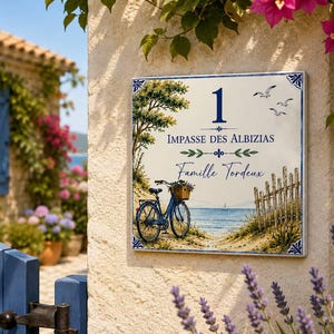 Puede incluir: Un letrero de casa cuadrado de cerámica con el número 1, el texto Impasse des Albizias y Famille Tordeux. La ilustración muestra una bicicleta azul con una cesta, una valla de madera, un camino de arena, el mar y tres aves volando.
