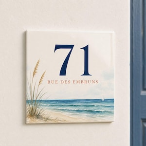 Könnte beinhalten: Ein quadratisches Hausnummernschild aus Keramik mit der Zahl 71 in Dunkelblau und dem Text RUE DES EMBRUNS in Orange. Das Design zeigt eine Küstenlandschaft mit Strandgras im Vordergrund und einem Segelboot auf dem Ozean.