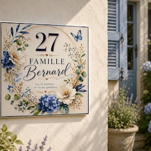 Op de afbeelding: Een vierkant huisbord met een crèmekleurige achtergrond met het nummer 27, de tekst FAMILLE Bernard en de zin Ici, le bonheur se vit au quotidien. Het ontwerp bevat een bloemenkrans met blauwe hortensia's, witte bloemen en een vlinder.