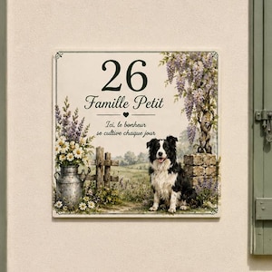 Könnte beinhalten: Ein quadratisches Hausschild mit der Nummer 26, dem Text Famille Petit und dem Satz Ici, le bonheur se cultive chaque jour. Das Design zeigt einen schwarz-weißen Hund, eine metallene Milchkanne mit Blumen und hängende Glyzinienranken.