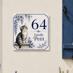 Könnte beinhalten: Ein quadratisches weißes Hausschild mit einer blauen Illustration eines getigerten Kätzchens, das auf einem Steinsims sitzt und von blauen Blumenranken umgeben ist. Das Schild zeigt die Zahl 64 in großer blauer Schrift über dem Text Famille Petit.