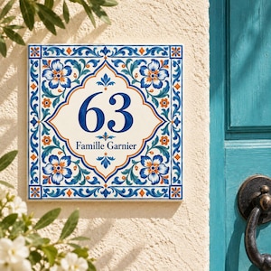 Könnte beinhalten: Ein quadratisches Hausnummernschild aus Keramik mit der Zahl 63 und dem Text Famille Garnier in Dunkelblau. Das Design zeigt ein weißes Zentrum mit einer dekorativen Bordüre aus blauen, türkisfarbenen und orangen Blumen auf cremefarbenem Grund.