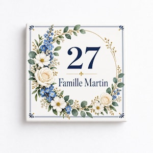 Könnte beinhalten: Ein quadratisches Hausschild mit weißem Hintergrund und dunkelblauem Rand. In der Mitte sind die Zahl 27 und der Text Famille Martin in Dunkelblau abgebildet. Ein Blumenkranz aus weißen Rosen, blauen Blüten und grünen Blättern umrahmt den Text.