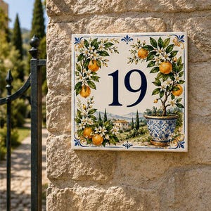 Puede incluir: Una placa de cerámica cuadrada para número de casa que muestra el número 19 en fuente azul oscuro. El diseño presenta naranjos pintados a mano con hojas verdes, flores blancas y frutos naranjas, junto a una maceta con motivos azules y blancos.