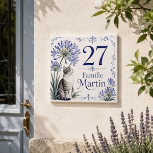 May include: A square white ceramic house sign featuring a blue painted illustration of a tabby cat sitting beneath blue agapanthus flowers. The sign displays the number 27 and the text Famille Martin in a dark blue serif font with decorative borders.