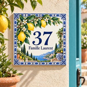 Puede incluir: Una placa de número de casa cuadrada de cerámica con el número 37 y el texto Famille Laurent en azul oscuro. El diseño incluye limones amarillos, hojas verdes, flores blancas y un paisaje costero mediterráneo con un borde azul.