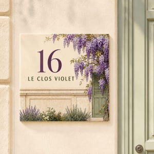 Op de afbeelding: Een vierkant huisnummerbord met het getal 16 en de tekst LE CLOS VIOLET. Het ontwerp toont een crèmekleurige muur met een groen luik, paarse blauweregenbloemen die naar beneden hangen en groene lavendelplanten aan de onderkant.