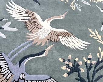 May include: A sage green area rug featuring a repeating pattern of flying cranes in shades of white, beige, and dark grey. The design includes stylised botanical elements, including slender reeds and dark leafy branches, integrated into the background.