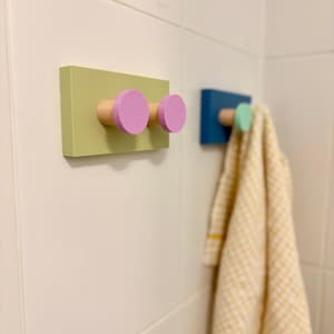 May include: Two rectangular wall-mounted hooks. The first is a light green base with two light pink circular pegs. The second is a dark blue base with a single light green circular peg, holding a yellow and white checkered towel.