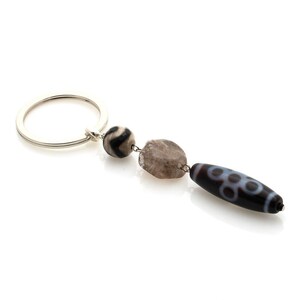 May include: A silver-toned metal keyring featuring three attached beads. The first is a small, round, black and white patterned bead, the second is a textured, irregular, grey stone bead, and the third is an elongated, dark brown bead with white circles.