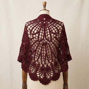 May include: A deep burgundy crochet shawl featuring an intricate, openwork spiderweb-inspired pattern that radiates from the center. The garment has a scalloped edge and is displayed on a mannequin, showcasing the detailed yarn work and texture.
